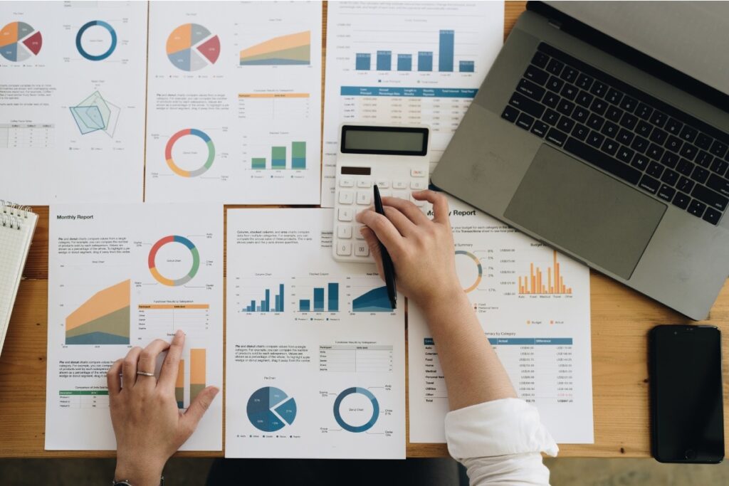 Overhead view of financial reports and charts spread across a desk with a hand using a calculator