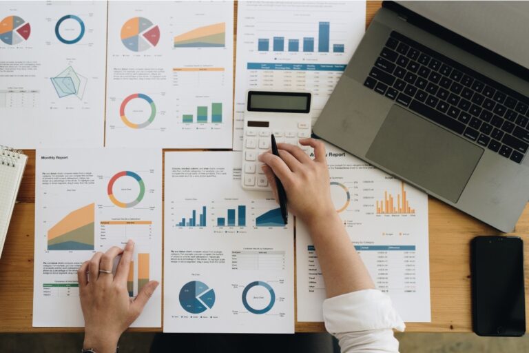 Overhead view of financial reports and charts spread across a desk with a hand using a calculator